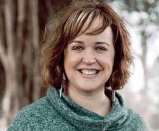 Woman with brown hair and dimples smiles, wearing a green sweater outdoors near trees.