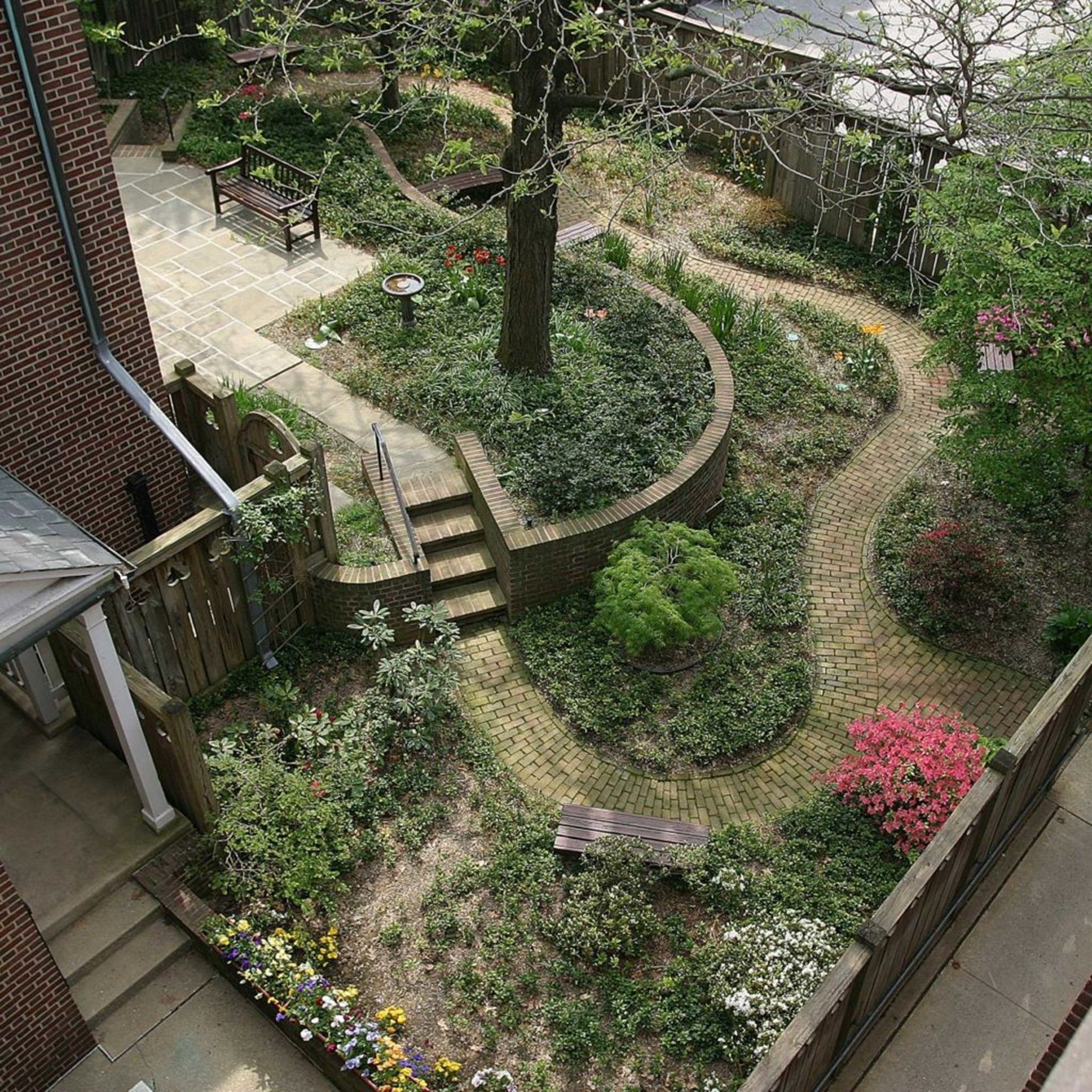 High-angle view of a small garden with winding brick paths, flowerbeds, and a central tree. A brick building and wooden fence border the space.