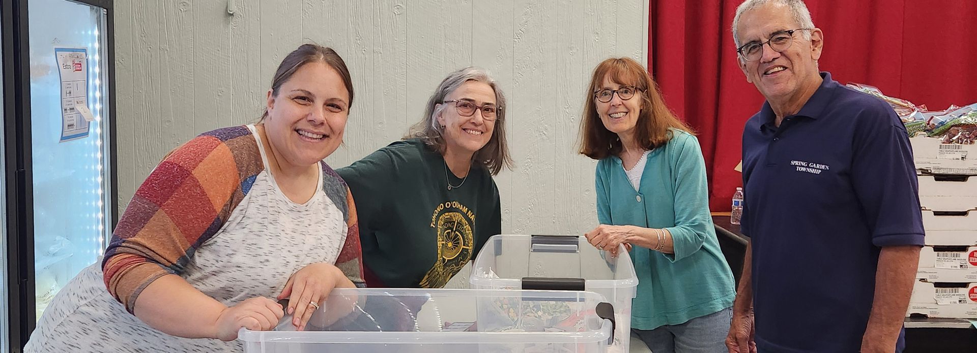 Four smiling people gather around a clear plastic container. Two women wear glasses, and the man on the right has short gray hair. They appear to be indoors near a red curtain.