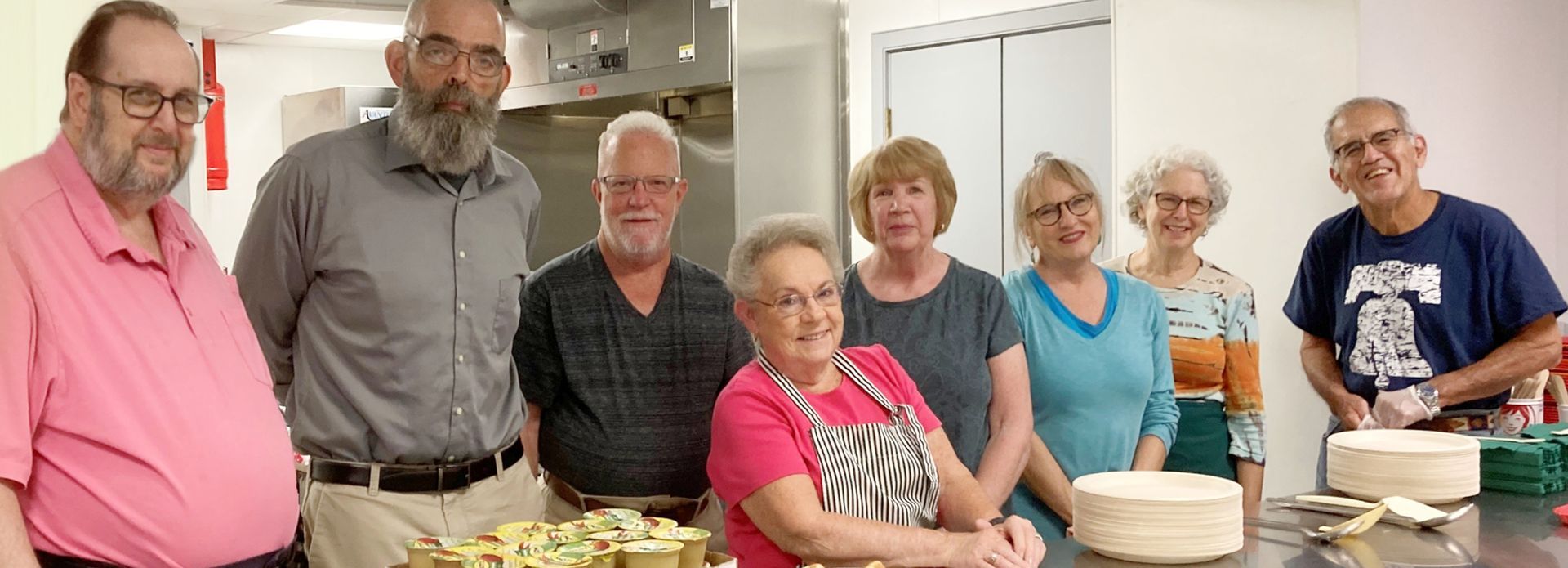 A group of people, both men and women, stand together in a kitchen, some smiling. They are gathered around a table with food.