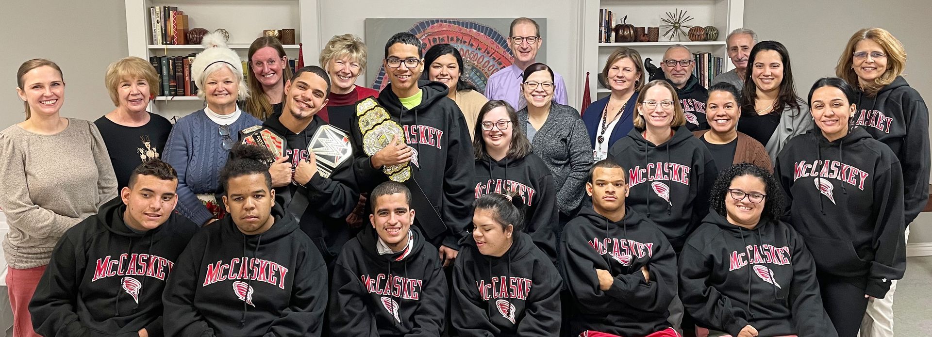 Group photo of people, mostly wearing black sweatshirts with a logo. Some are smiling, posing in a room with shelves and artwork.