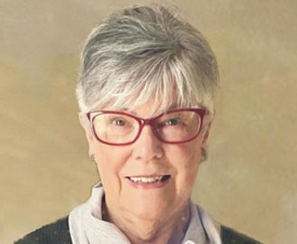 Woman with short gray hair and glasses, smiling. Wearing red-rimmed glasses, a gray sweater, and scarf.