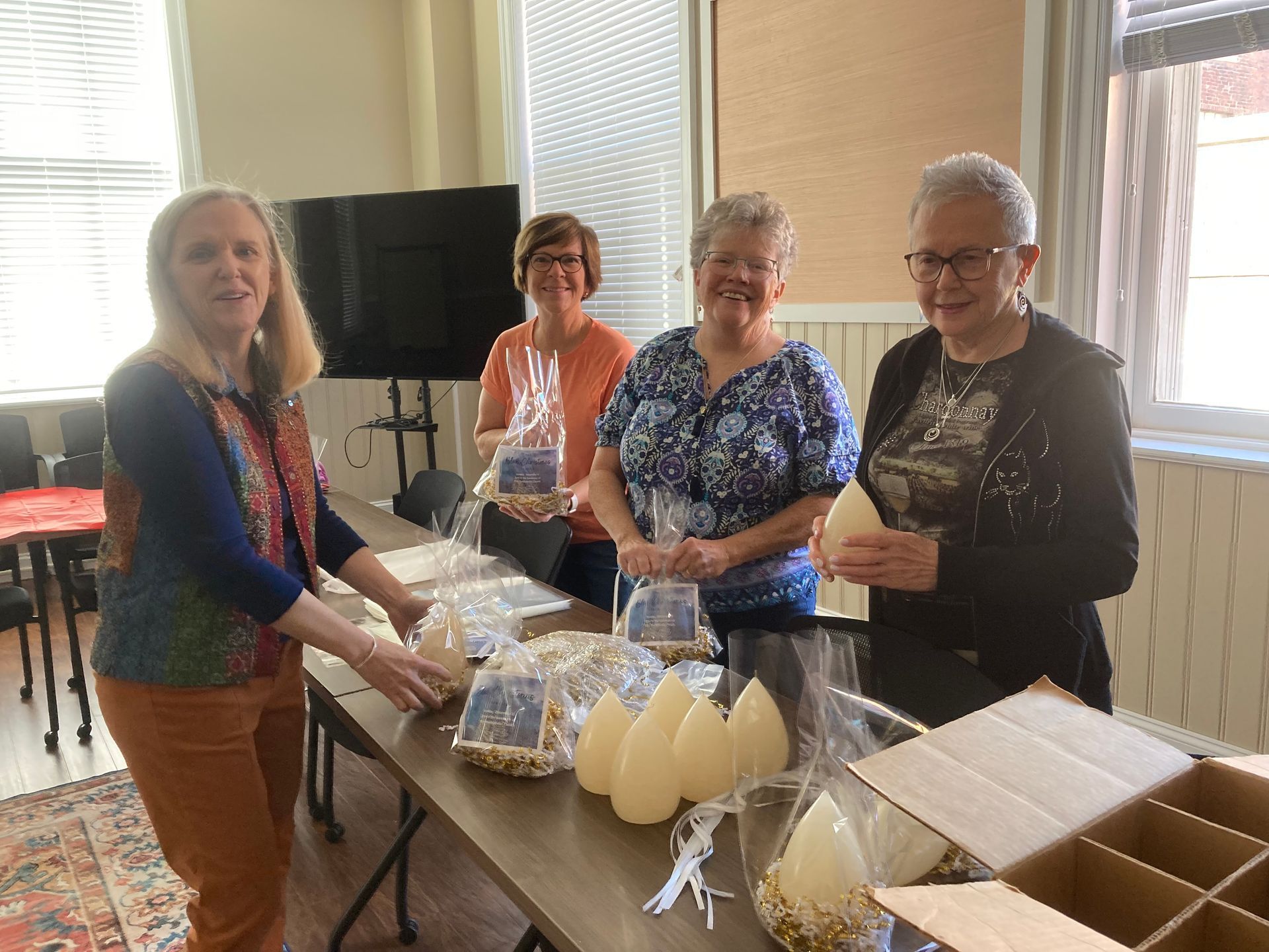 Four women standing around a table, arranging candle-shaped items, presumably for a fundraiser. Two women smile, and the setting appears to be a church hall.