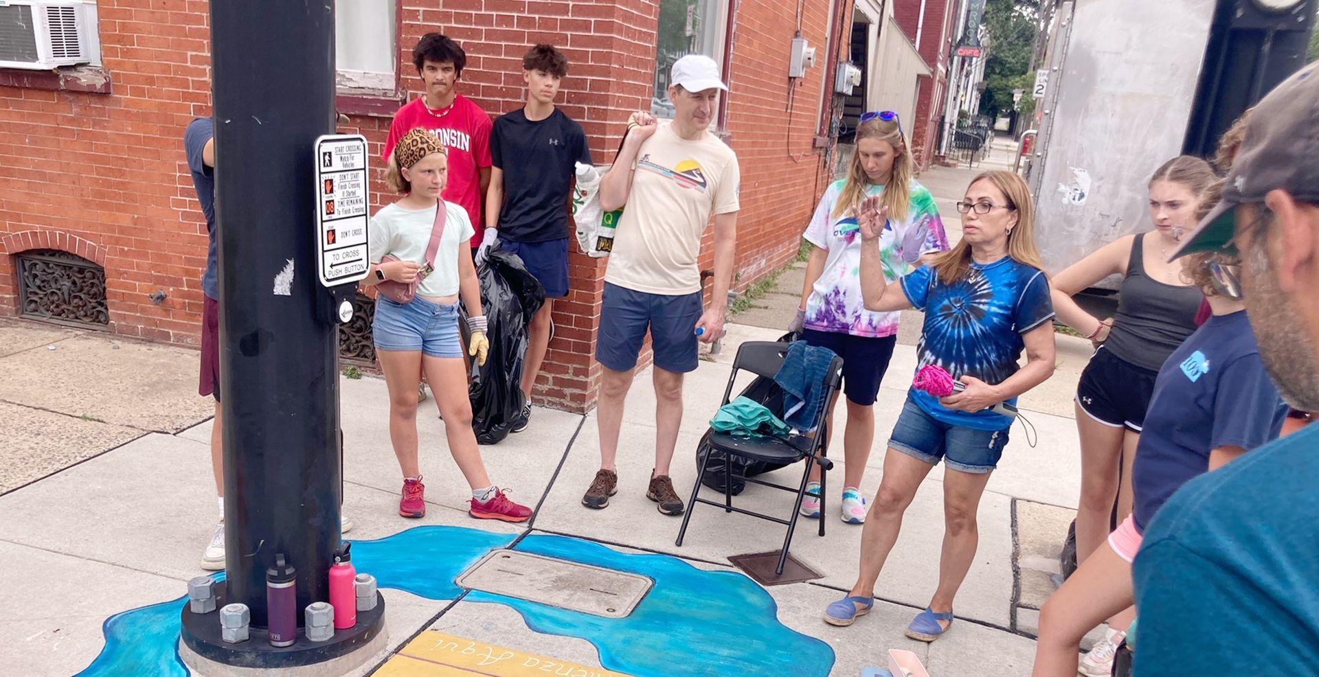 A group of people, mostly teenagers, stand around a painted sidewalk scene. They appear to be listening to a woman who is gesturing with her hands.