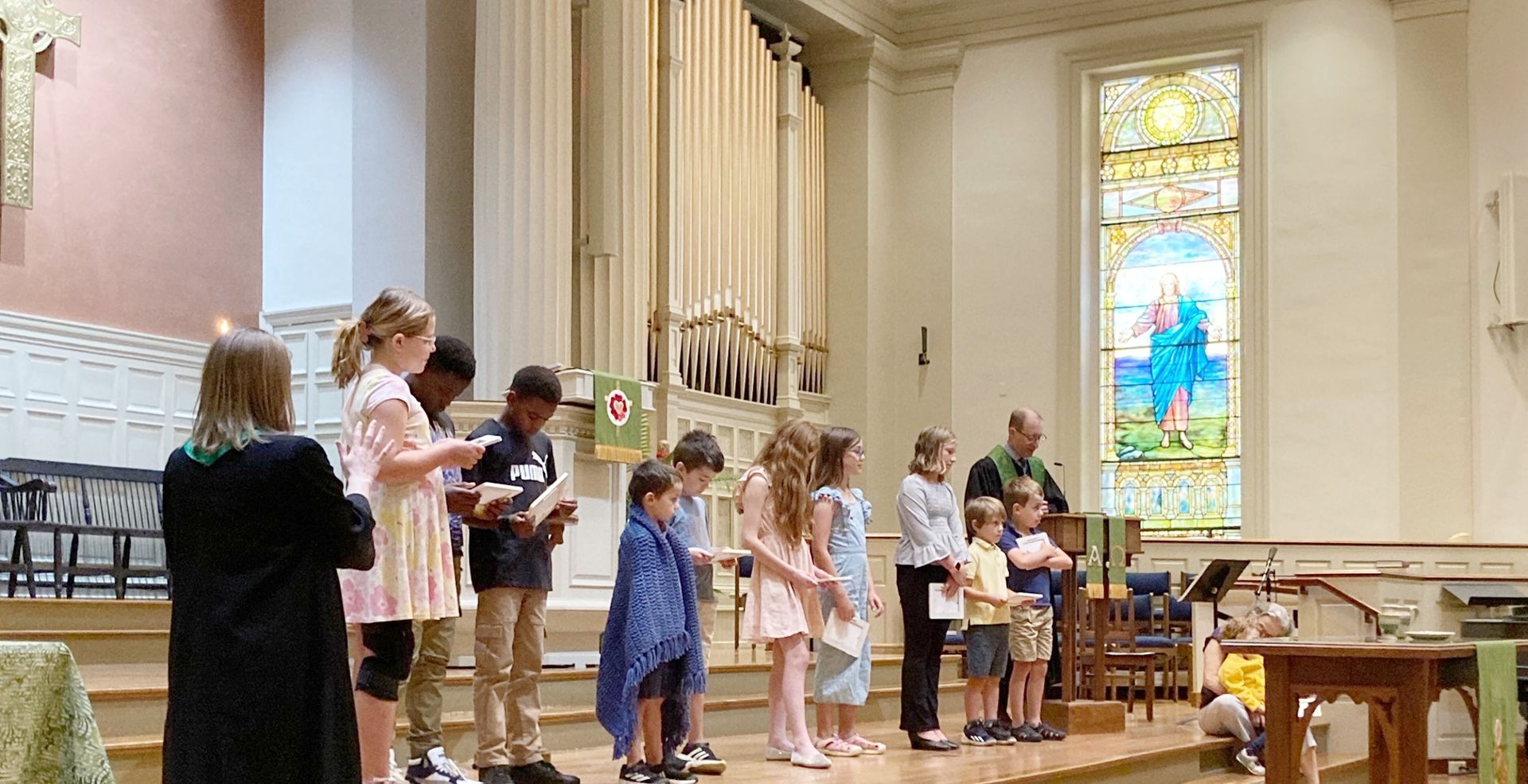 Children stand on a stage in a church, holding books and listening. A person in a green robe leads them, with a stained-glass window in the background.