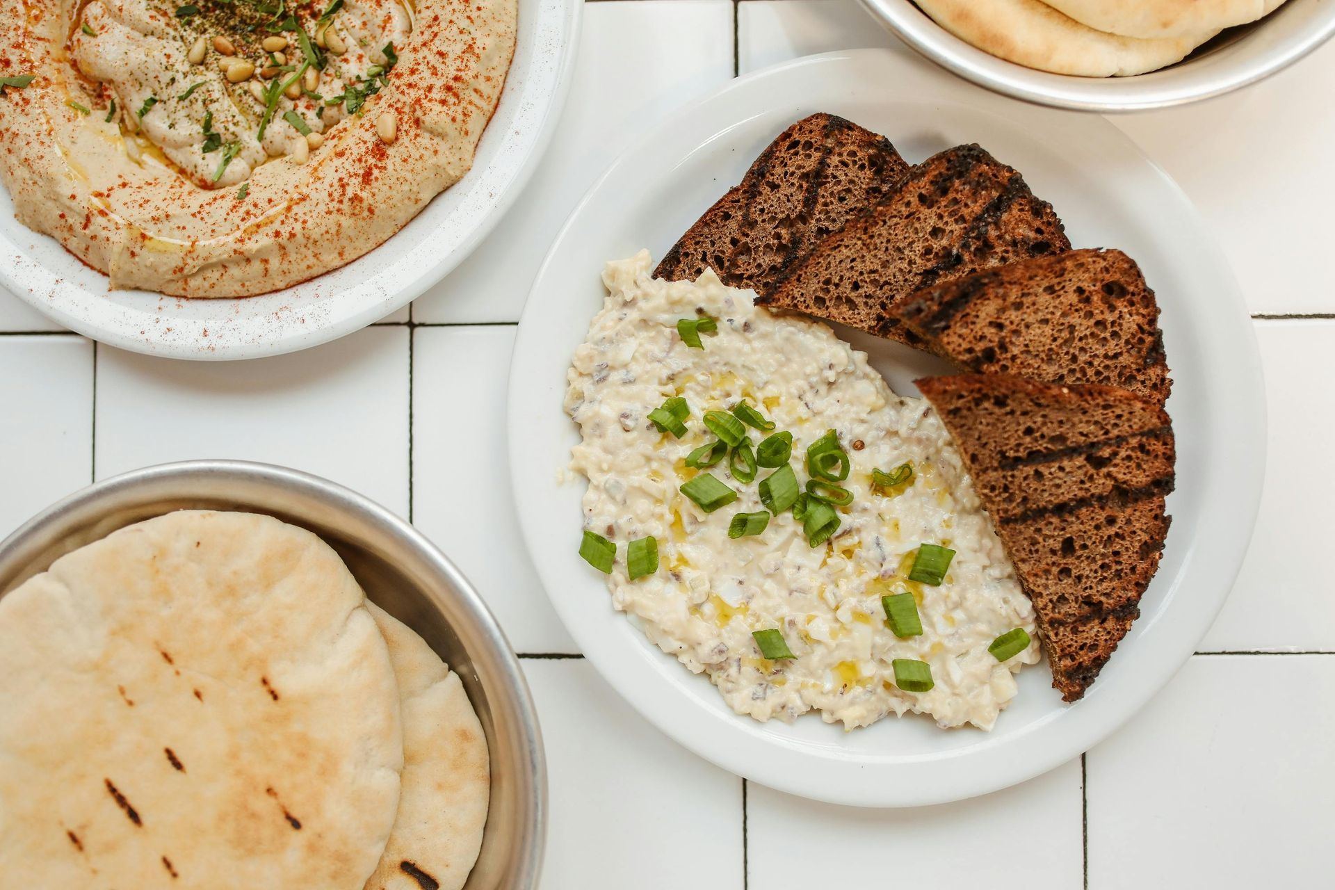 Overhead view of Middle Eastern dishes: hummus, baba ghanoush, pita bread, and grilled dark bread on a white tiled surface.