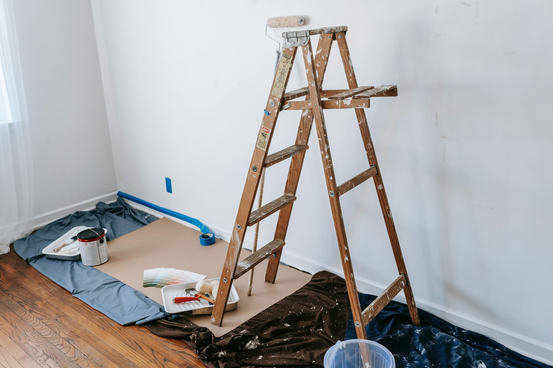 Ladder in a room with painting supplies, drop cloths, and taped baseboards during wall prep