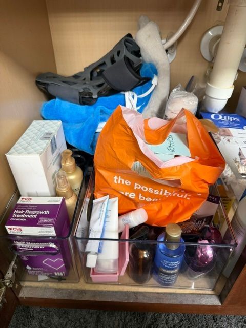 Cluttered bathroom shelf with assorted toiletries, tissue boxes, bags, and cleaning items inside a cabinet.