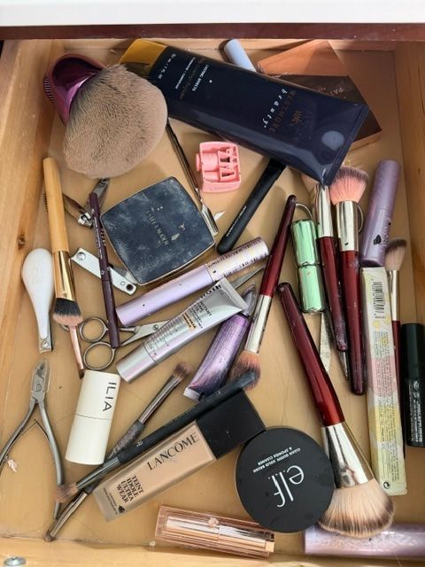 Scattered makeup products and brushes on a wooden surface, including palettes, tubes, pencils, and a compact.