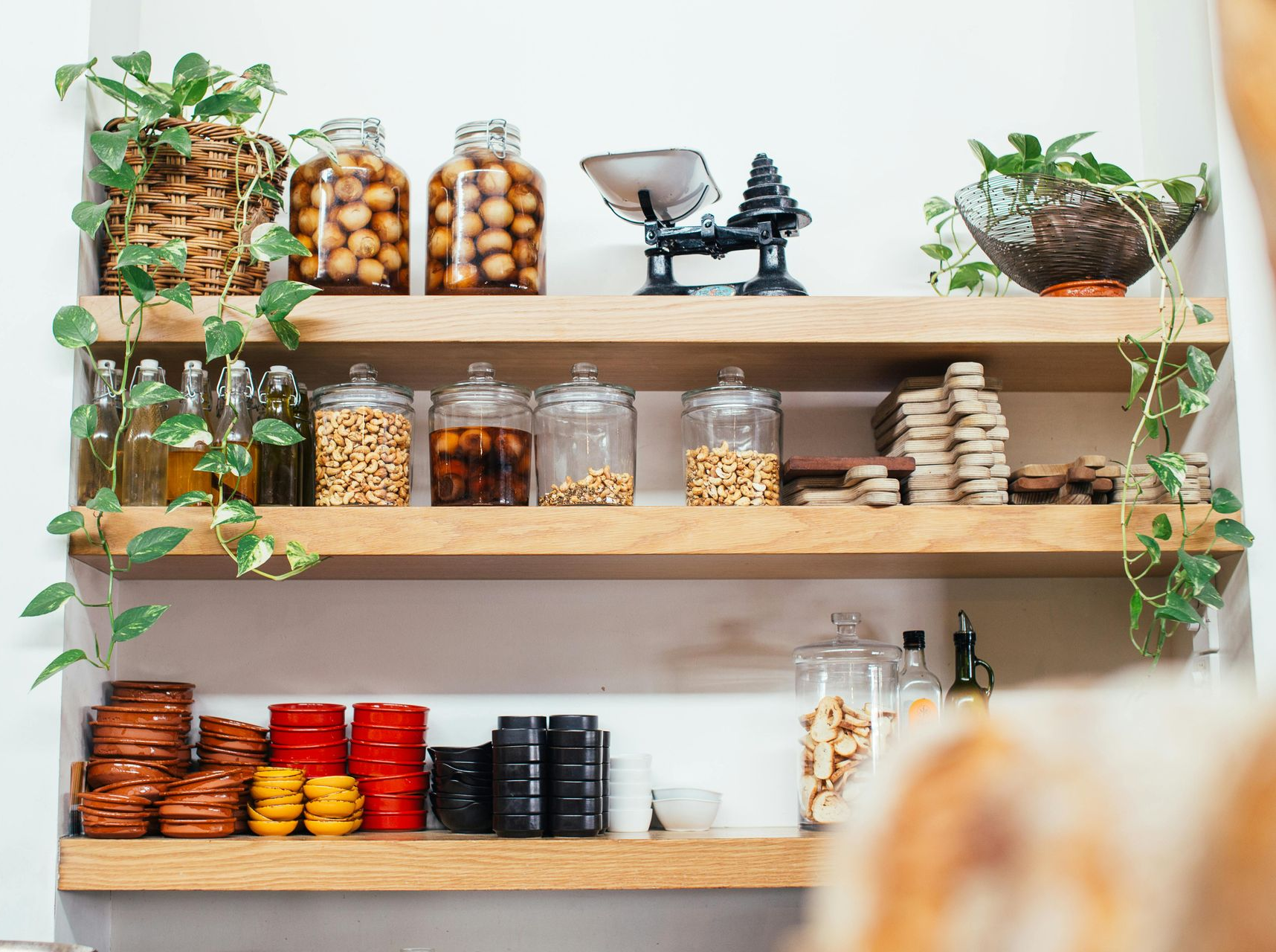 Wooden shelves with glass jars of grains, dried goods, plants, and a vintage scale in a bright kitchen.