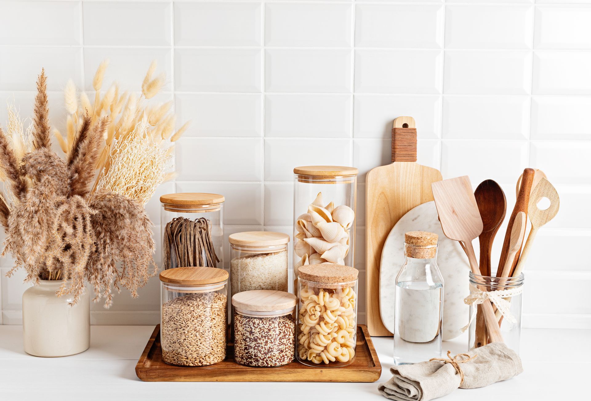 Neutral-toned kitchen shelf with glass jars of grains, dried flowers, wooden utensils, and cutting boards