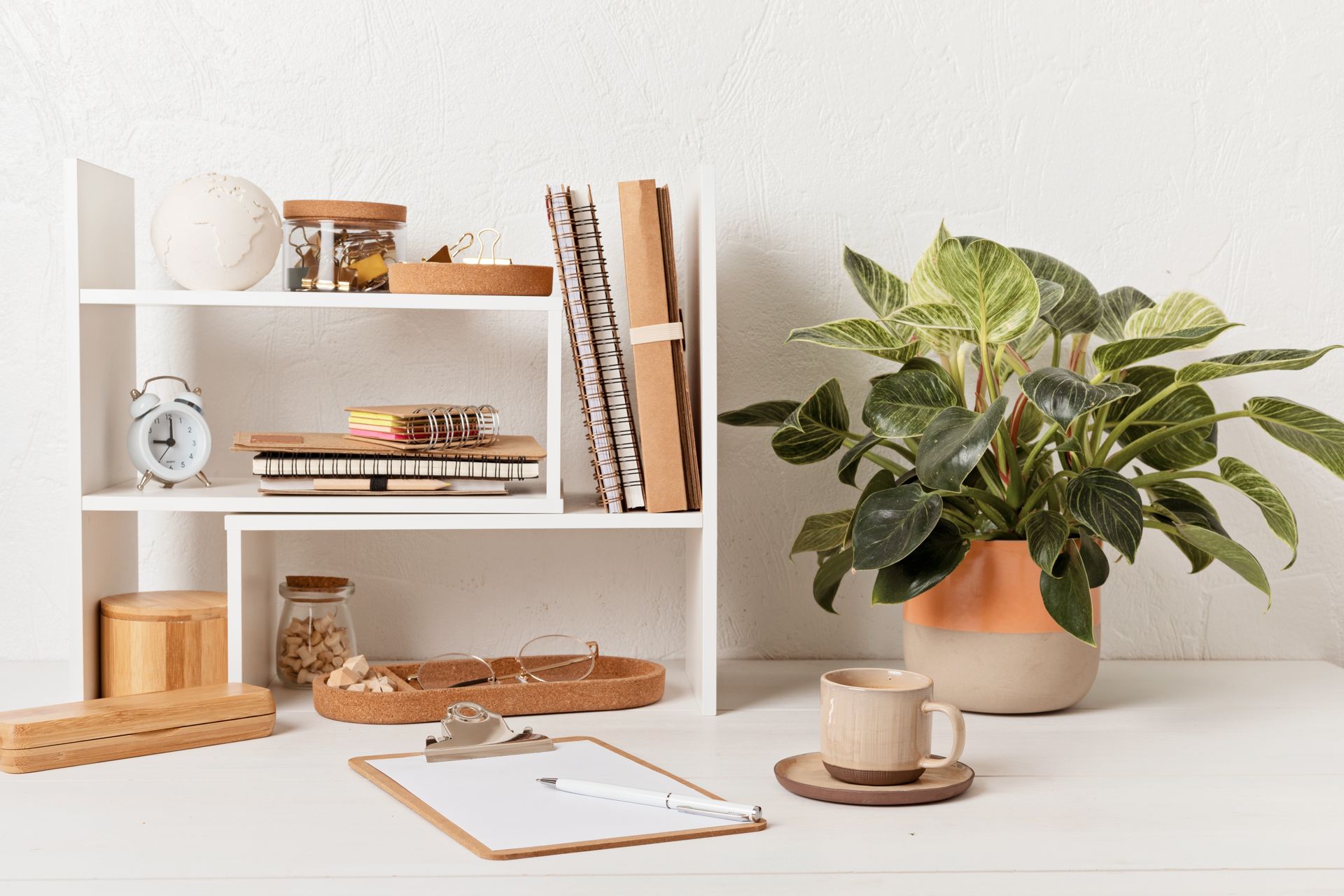 Minimalist desk with open notebook, plant, coffee cup, and white shelving with decor
