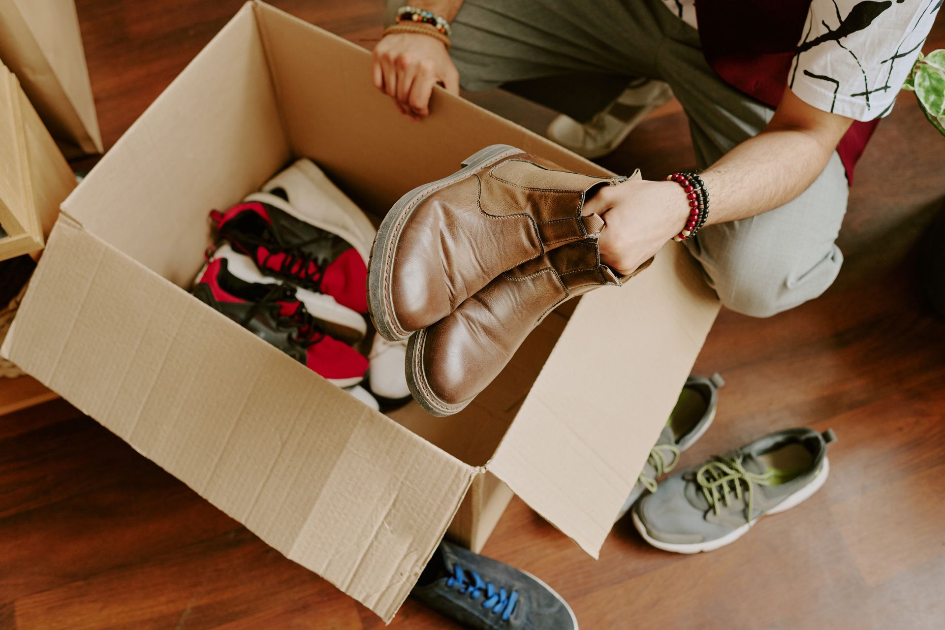Person packing shoes into a cardboard box on a wooden floor, with sneakers and a bag nearby