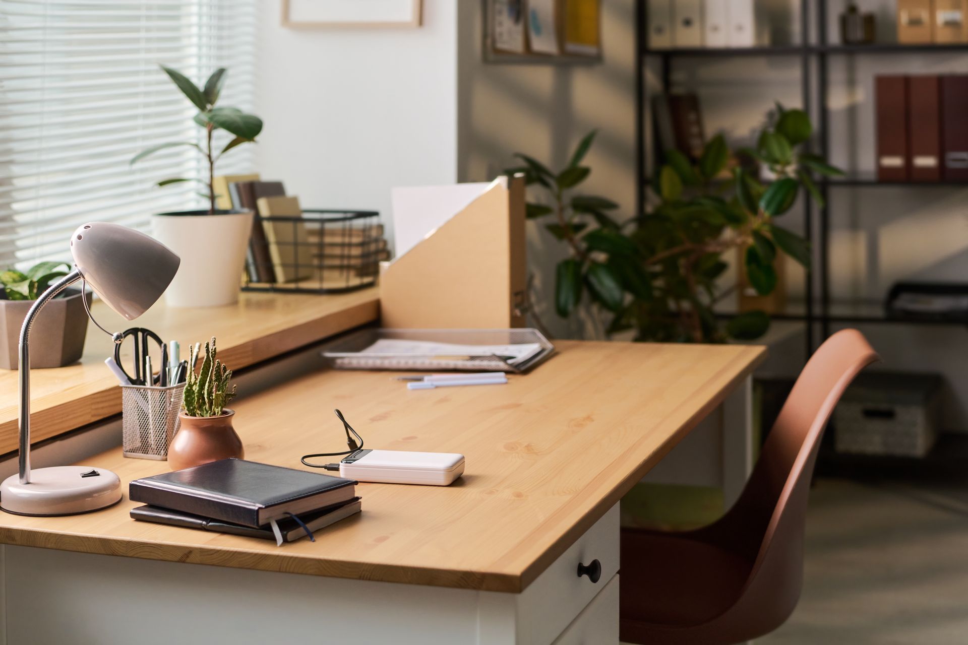 Modern office desk with lamp, notebook, chair, and potted plants by a window