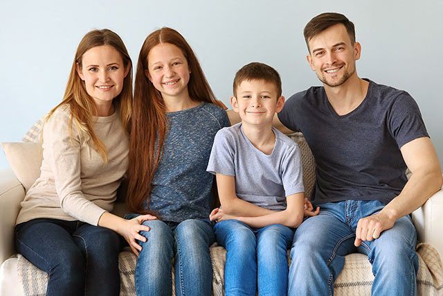Family of four sitting on a couch, smiling. Two children, two adults, light blue wall.