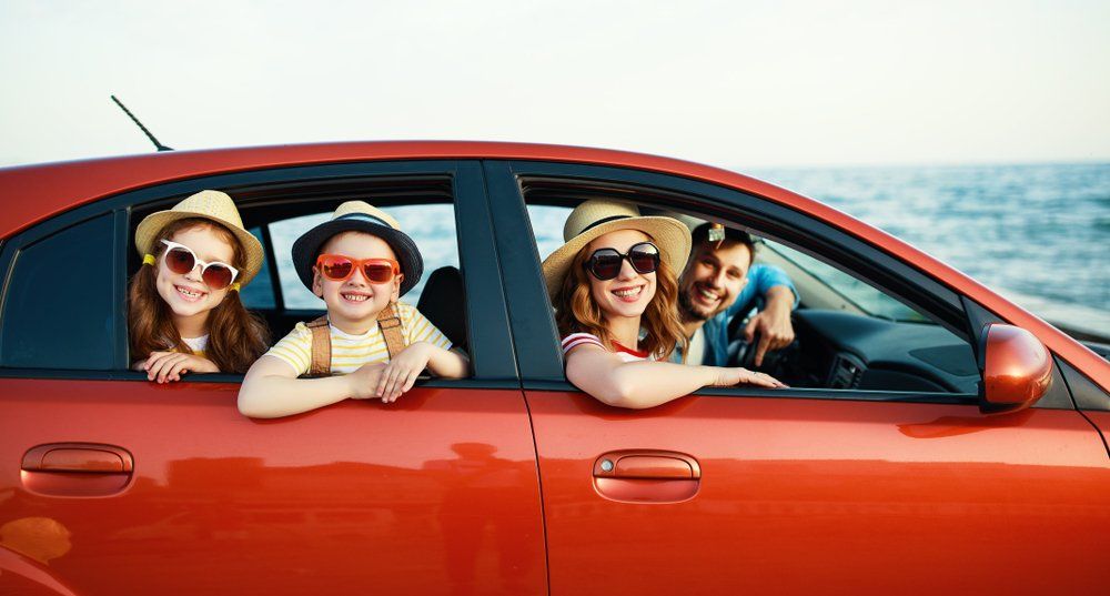 Famille dans une voiture orange à la plage, souriante et portant des lunettes de soleil et des chapeaux.
