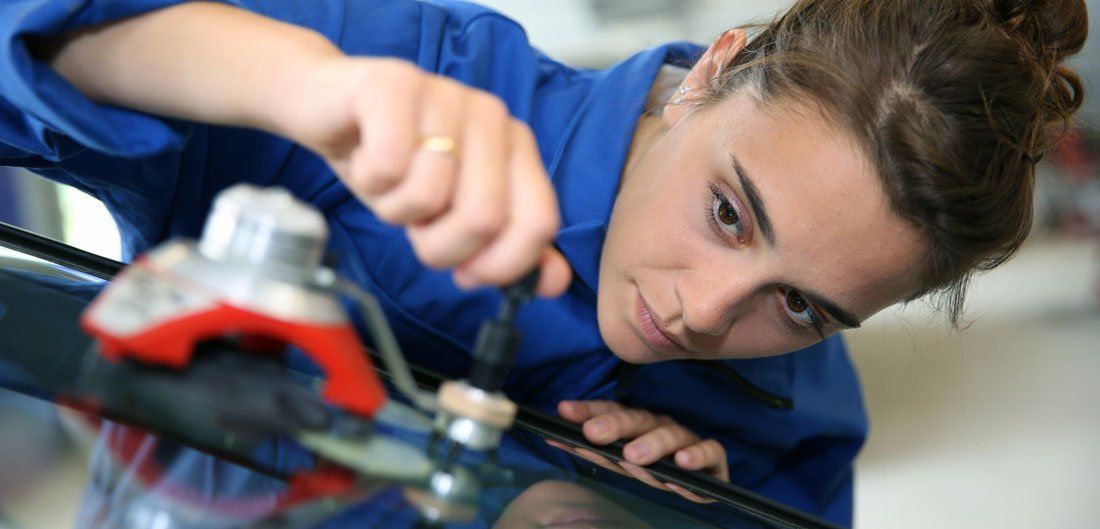 Une femme en salopette bleue répare un pare-brise, intensément concentrée.