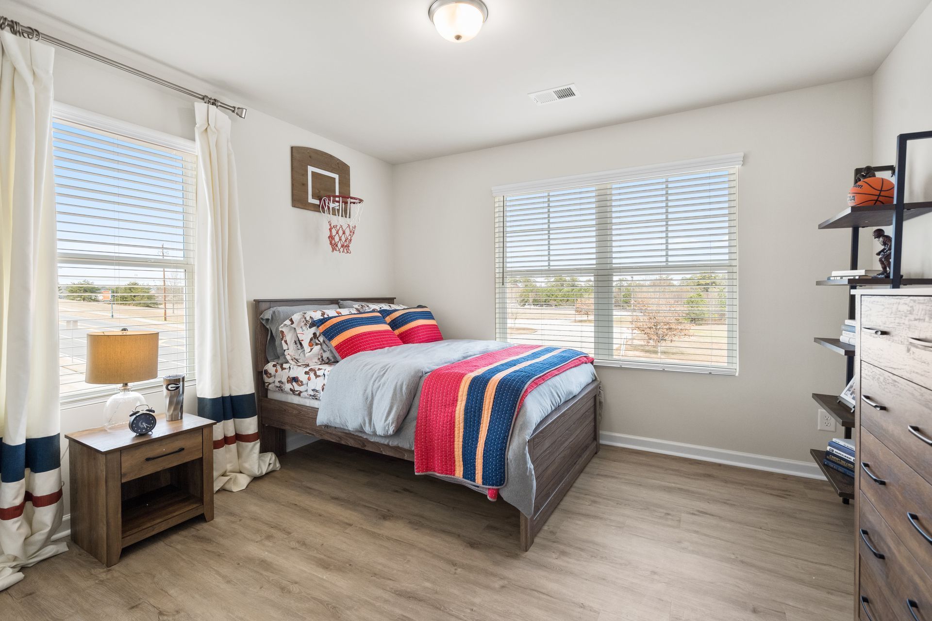 A bedroom with a bed , nightstand , dresser and two windows.