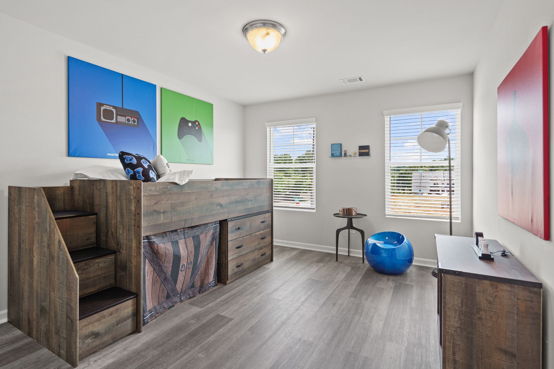 A bedroom with a bunk bed , dresser , chair and paintings on the wall.