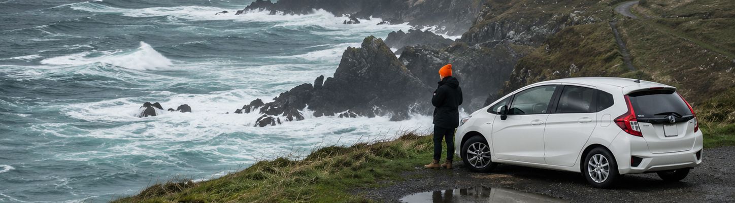 Person standing by a white SUV on a windy rocky coastline with crashing waves