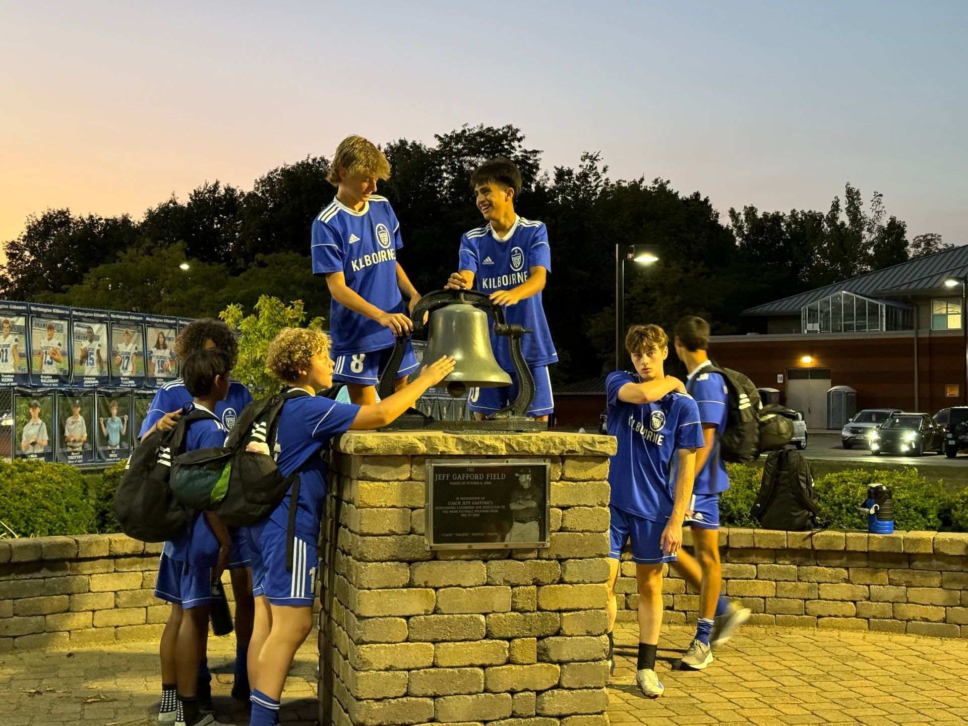 Soccer team in blue uniforms rings a large bell on a brick pedestal at dusk.