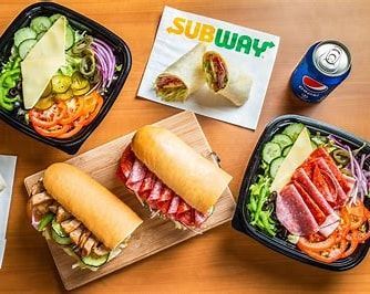 Subway sandwiches, salad bowls, and wrap on a wooden table with a Pepsi can and Subway logo.