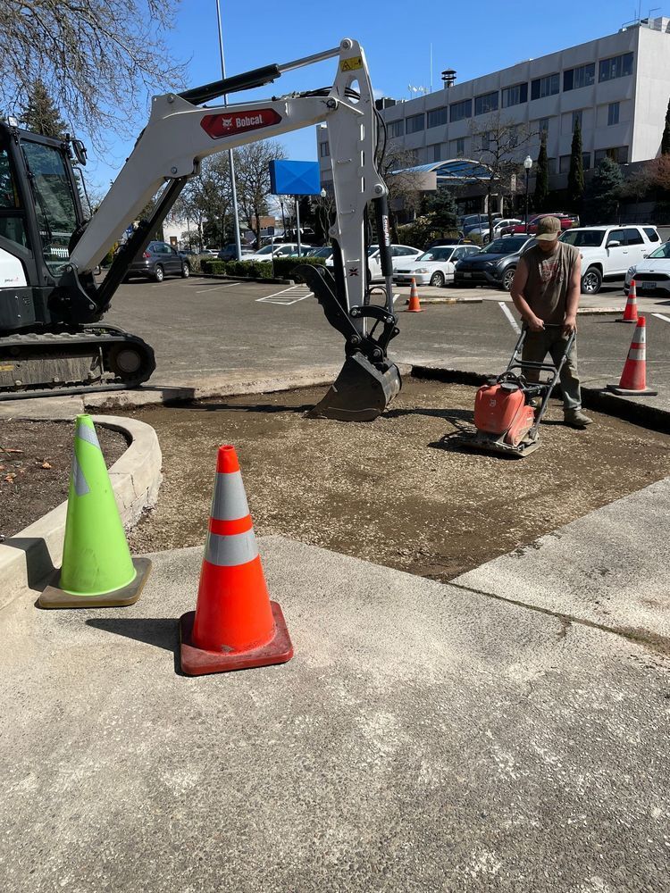 A man is working on a sidewalk next to a bulldozer and traffic cones.