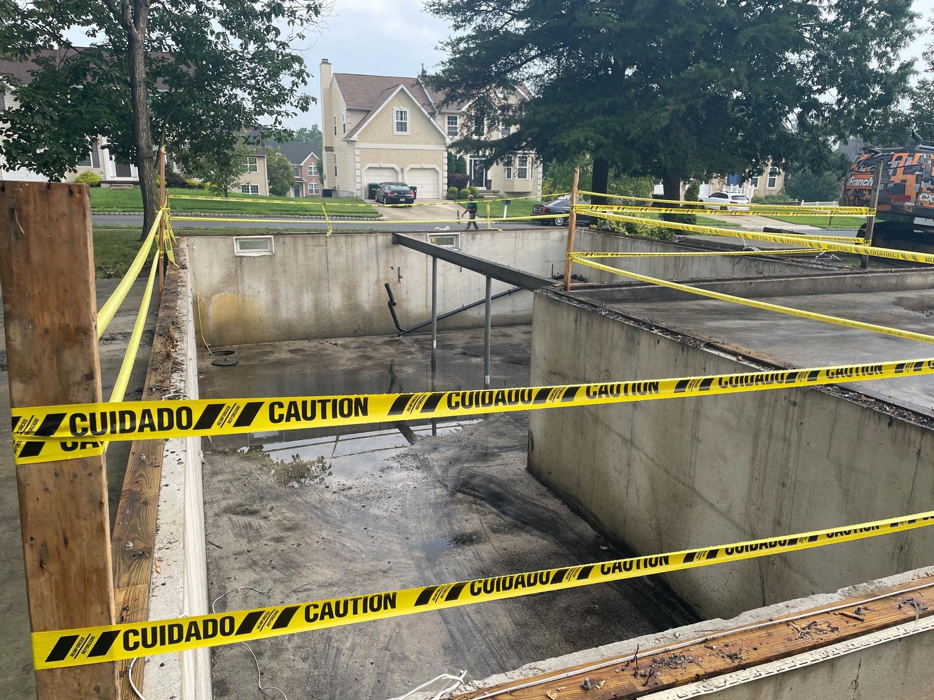 Construction site with concrete foundation, caution tape, and a house in the background.