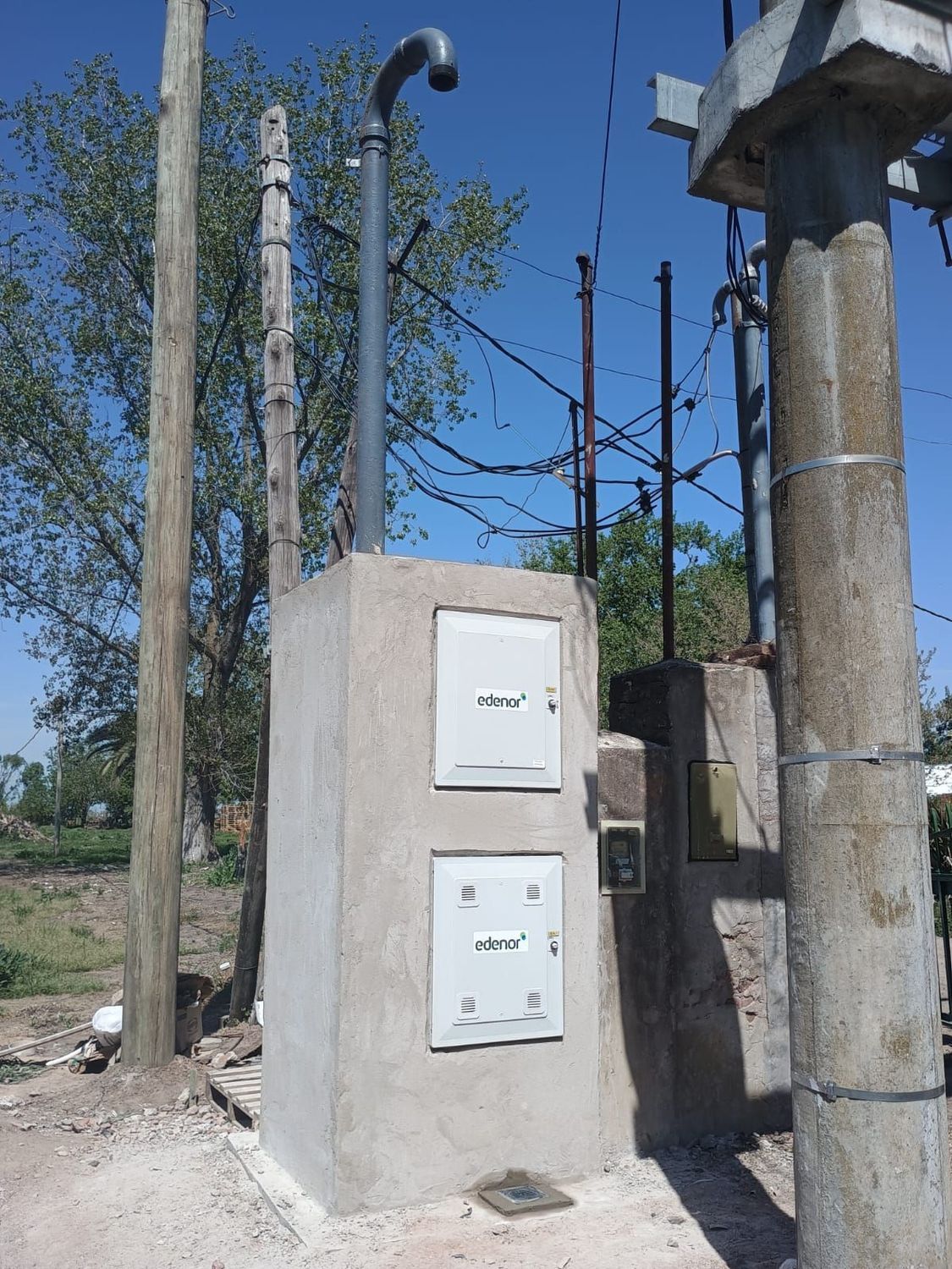 Un armario eléctrico con dos puertas blancas empotrado en un pilar de hormigón, con un conducto y postes de servicios públicos cerca.