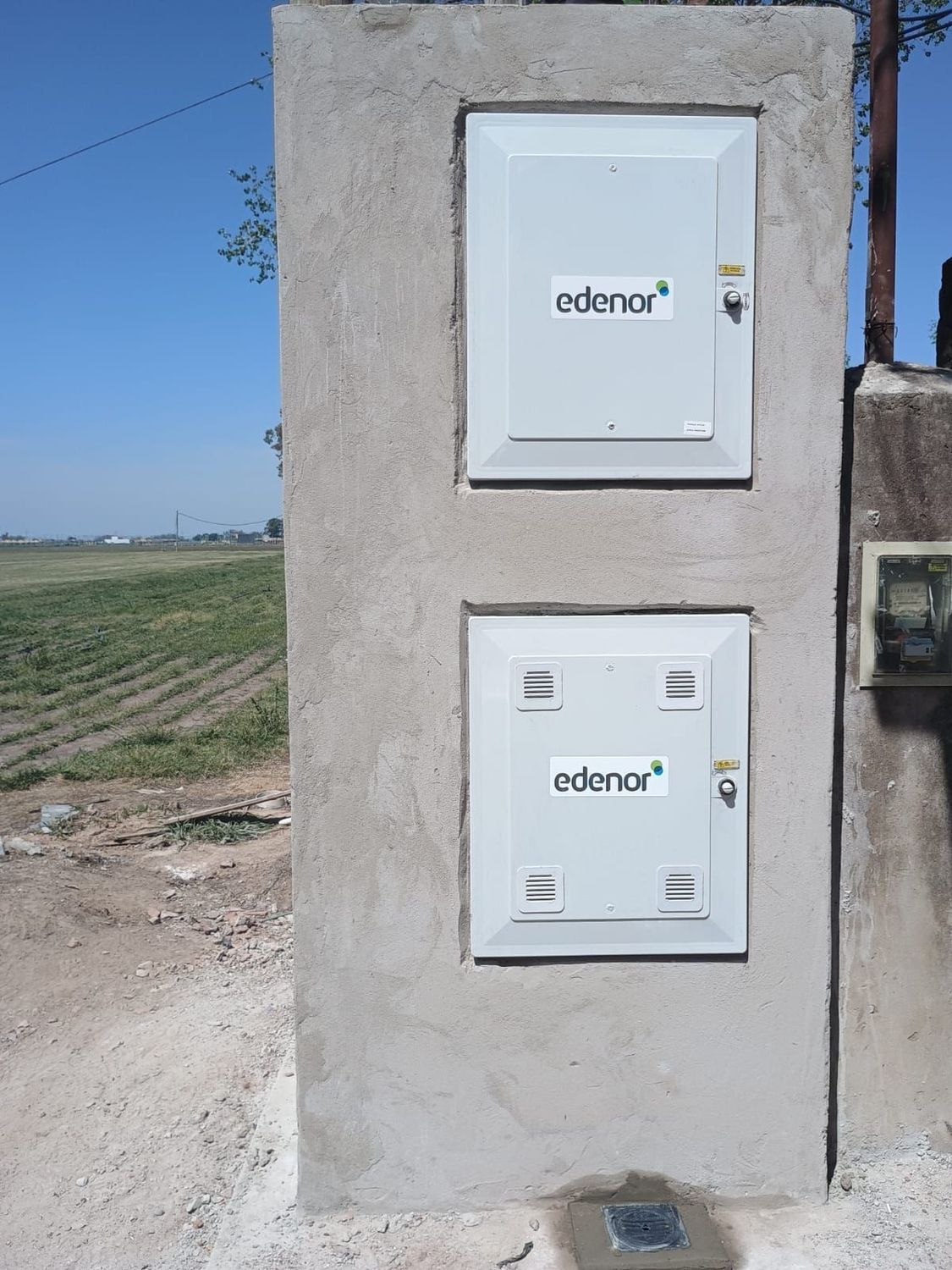 Un armario eléctrico con dos puertas blancas empotrado en un pilar de hormigón, con un conducto y postes de servicios públicos cerca.