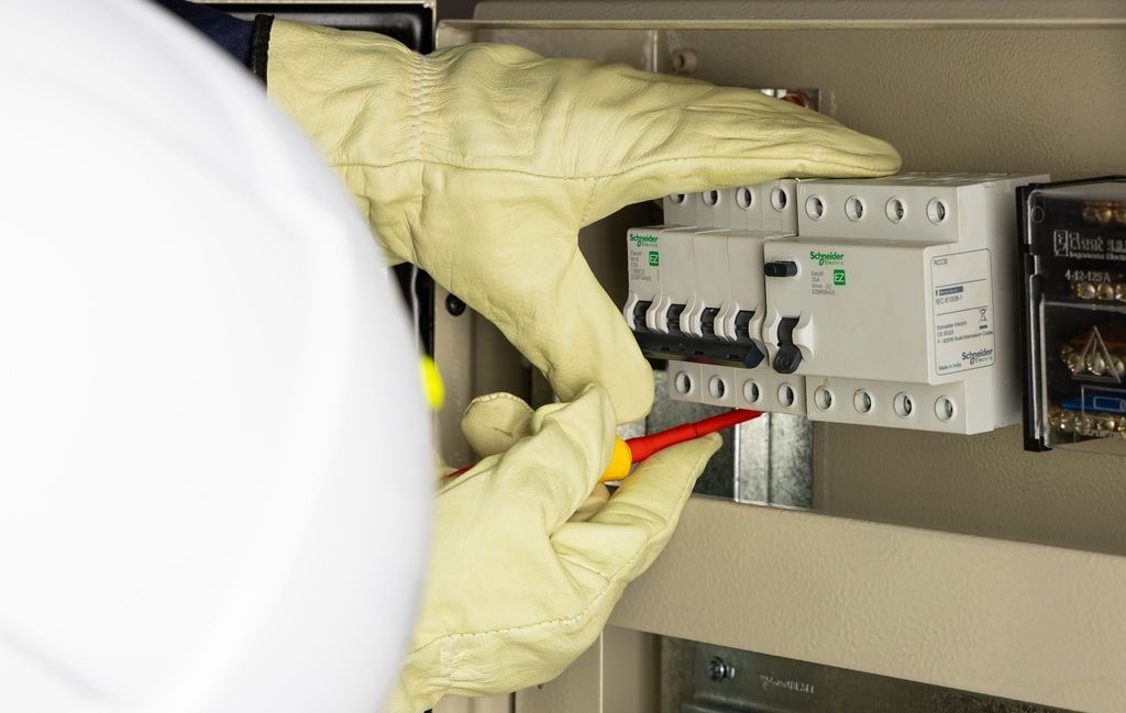 Una persona con guantes protectores utiliza un destornillador para trabajar en un panel de interruptores eléctricos blanco dentro de un armario.