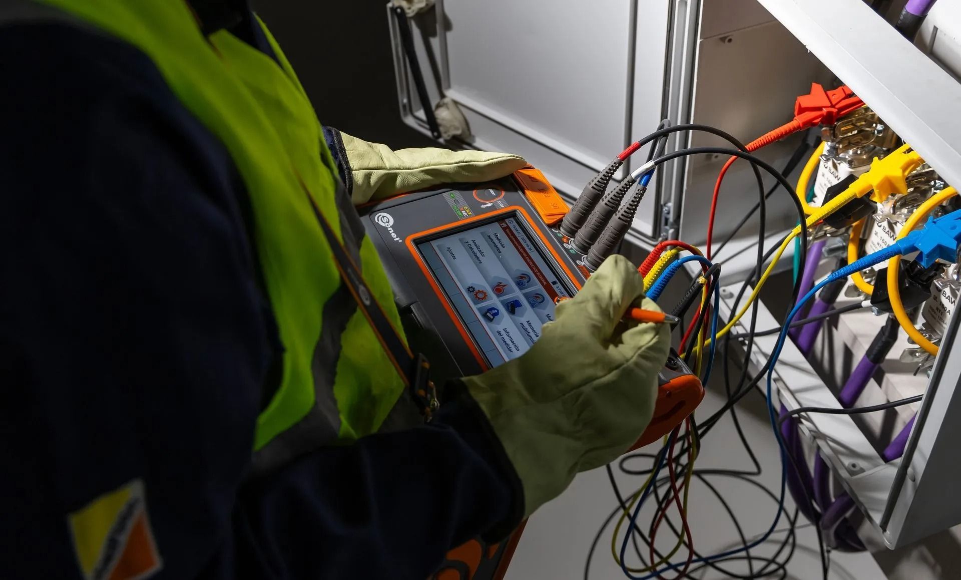 Un técnico, con chaleco de seguridad y guantes, utiliza un dispositivo de diagnóstico portátil para probar el cableado complejo de una caja eléctrica.