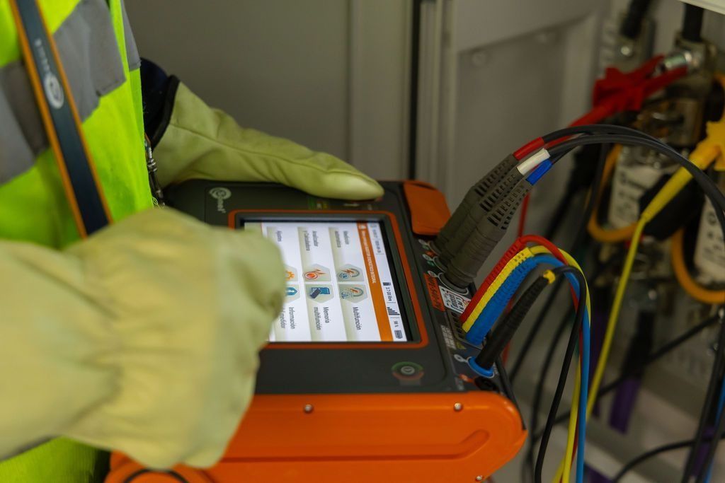 Un técnico con un chaleco reflectante y guantes aislantes utiliza un comprobador electrónico portátil en el cableado eléctrico.