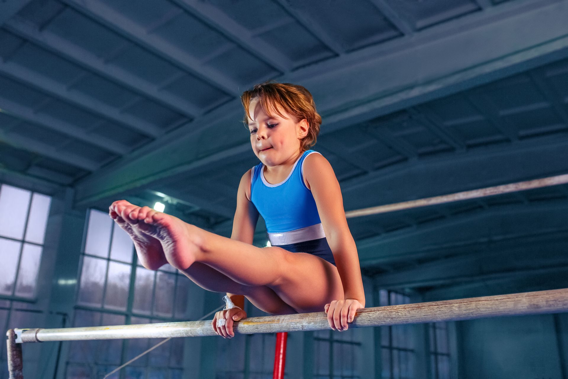 A young girl is holding an L-SIT on a horizontal bar in a gym.