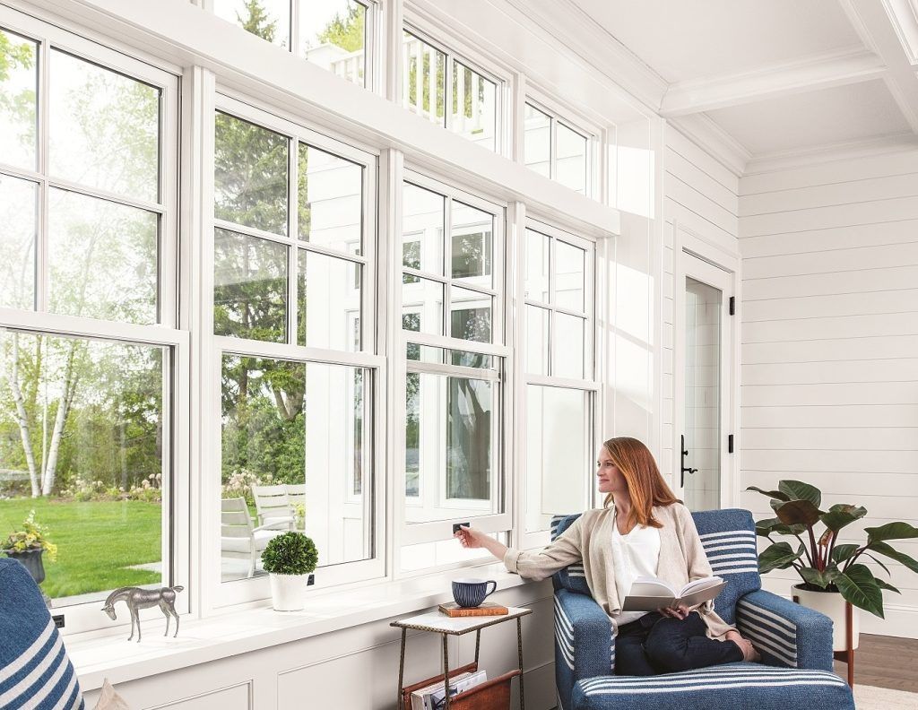 Woman seated in a light-filled room with large windows, adjusting a remote control. She is holding a tablet, a plant and small table are nearby.