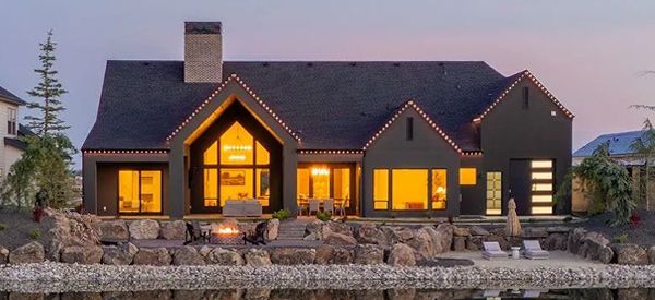Dark modern house with glowing windows, chimney, and stone accents, set near a lake at dusk.