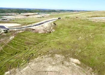 Aerial view of a hilly, grassy landscape with a road and vehicles. Green and brown terrain, sunny day.