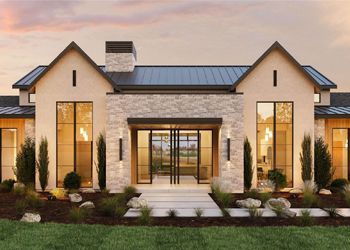 Modern home exterior with stone facade, large windows, and manicured landscaping under a sunset sky.