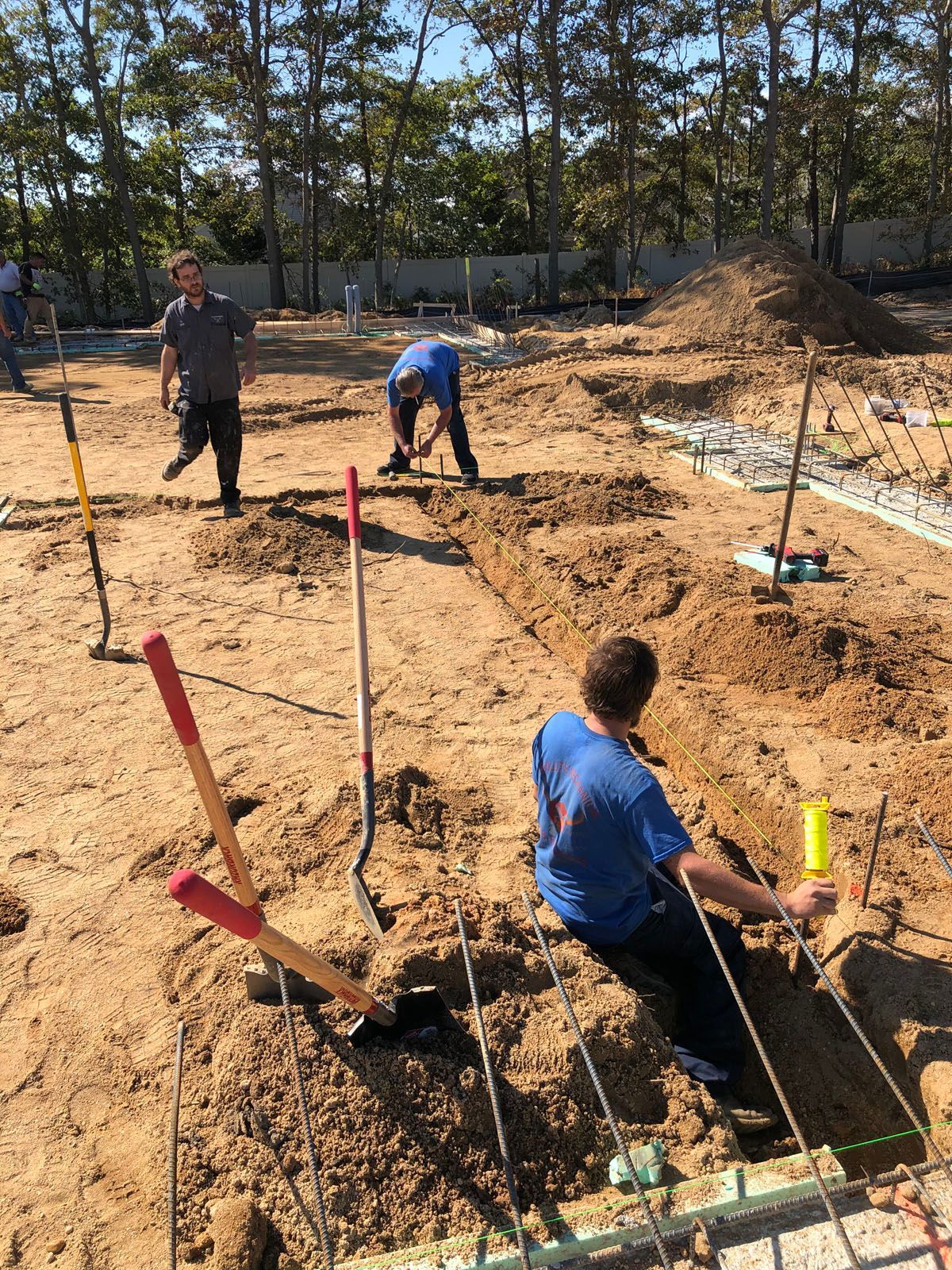 Three workers in a sandy, outdoor construction site dig trenches for a foundation among metal rebar and shovels.