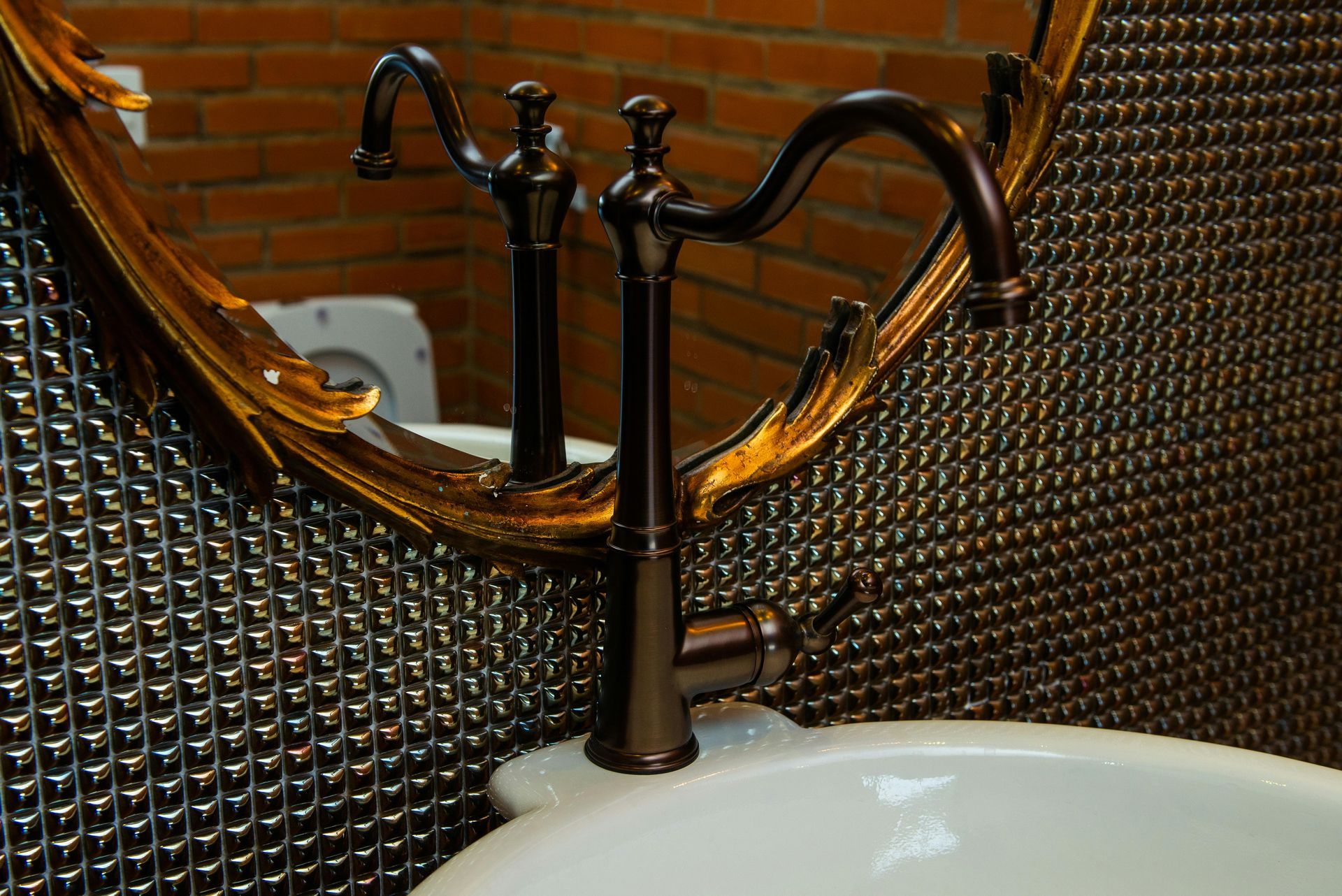 A vintage-style bronze faucet sits before a textured wall and an oval mirror reflecting a white toilet.