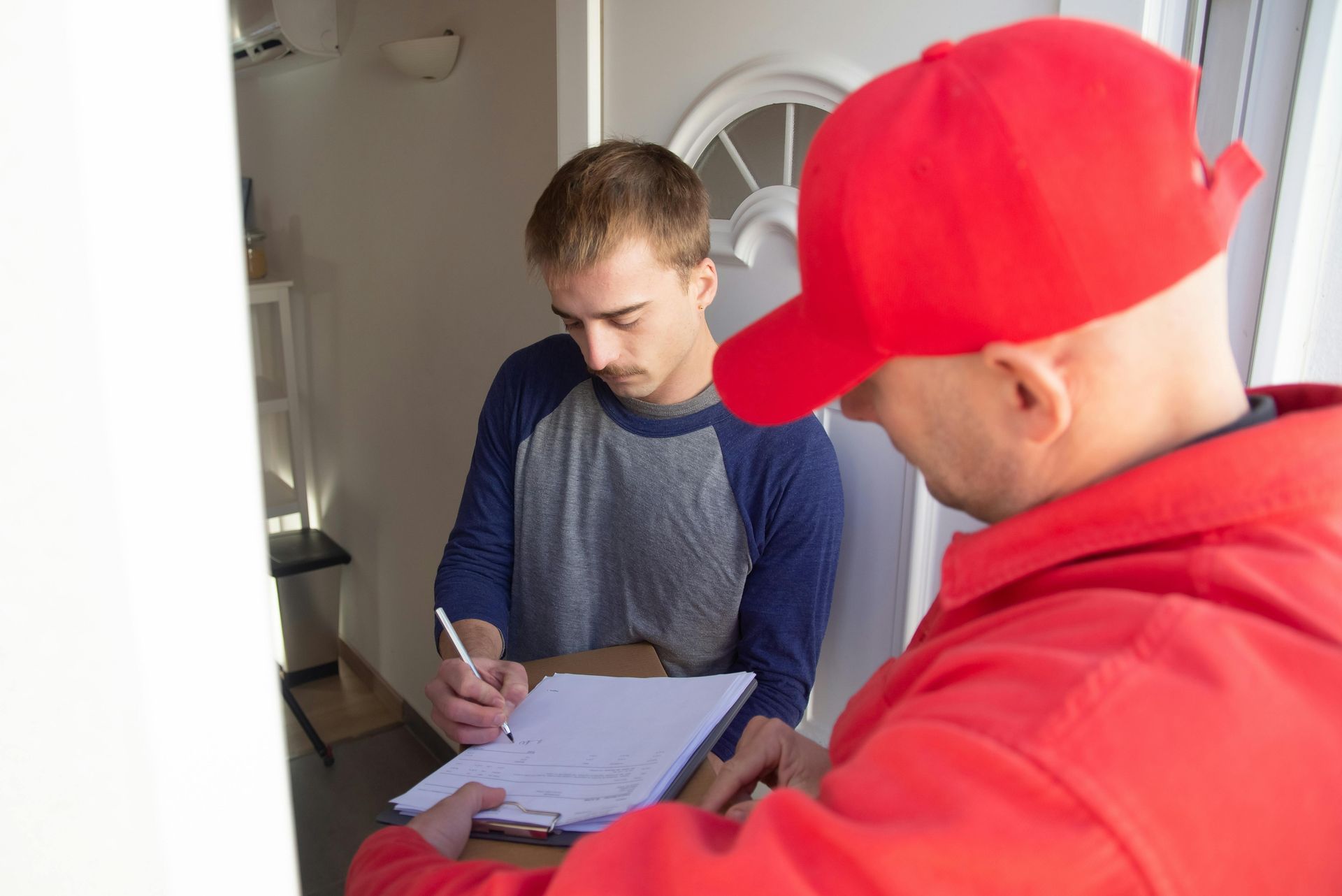 A person in a red uniform holds a clipboard for another person to sign while standing in a doorway.