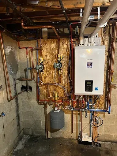 Wall-mounted boiler unit and copper plumbing setup with circulating pumps and a grey expansion tank in a basement.