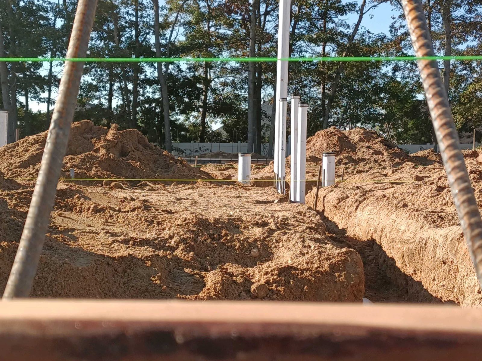 A view through vertical rebar poles at a construction site with trenches, dirt mounds, and white plumbing pipes.