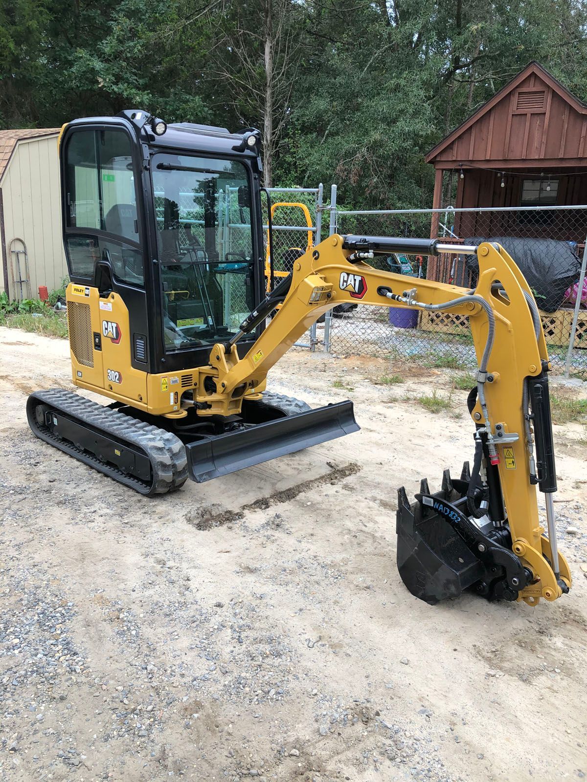 A yellow Caterpillar mini excavator sits on a dirt surface in front of a shed and chain-link fence.