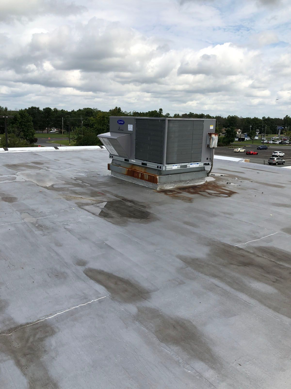 A gray HVAC unit sits on a flat, light-gray commercial roof under a cloudy sky, with a parking lot visible in the distance.
