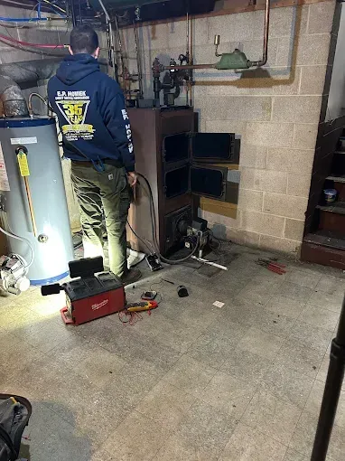 A technician inspects a furnace in a basement, using a red diagnostic tool on the concrete floor.