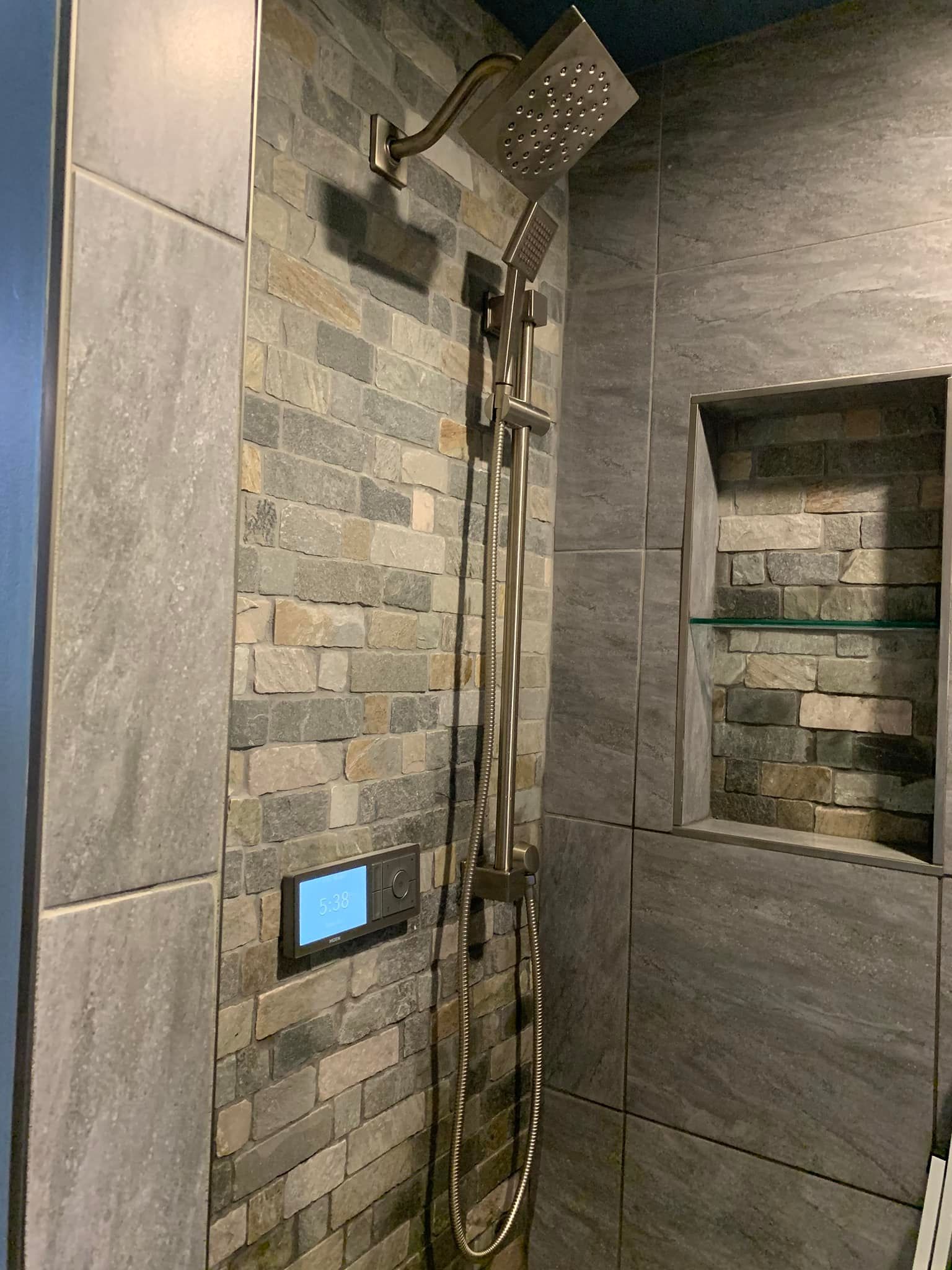 A modern shower with textured stone tile walls, a stainless steel rainfall showerhead, handheld sprayer, and digital control.