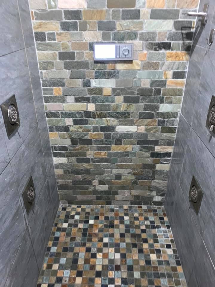A shower interior featuring gray slate tile walls, a decorative stone-patterned back wall, and a multi-colored mosaic floor.