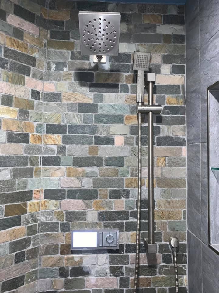 A modern shower with a square rainfall showerhead, handheld sprayer, and digital temperature control on a stone wall.