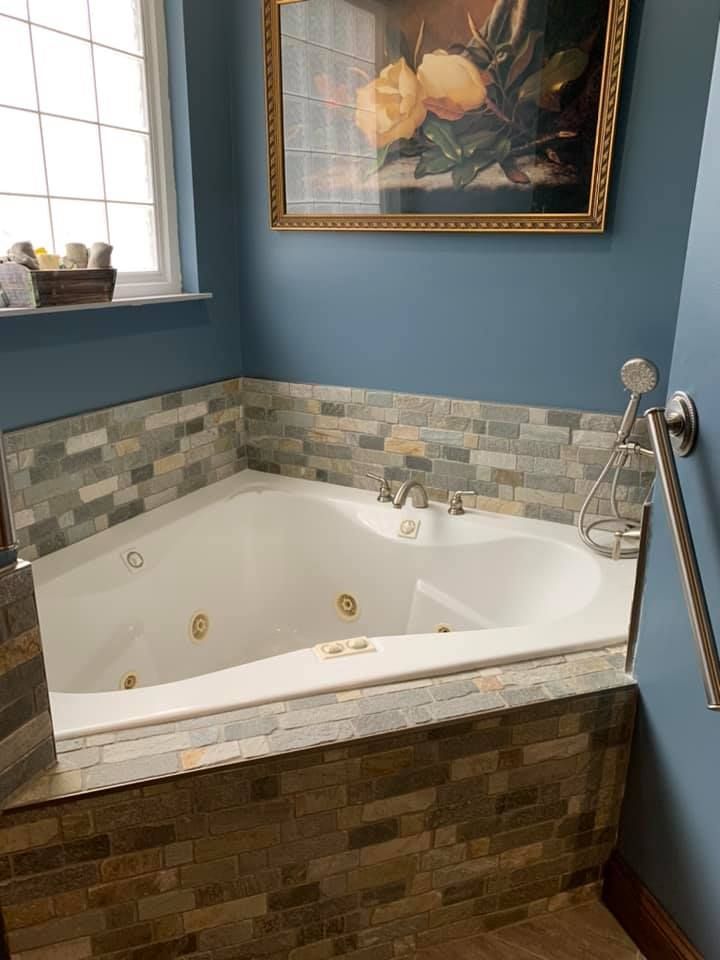A corner jetted bathtub with a multi-colored stone tile surround, set against blue walls under a floral painting.
